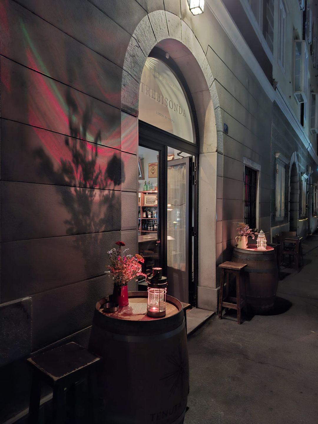 Ingresso di Trebisonda la sera: portale ad arco con la scritta sulla lunetta, porta aperta e botti-tavolino con lanterne accese sul marciapiede.
