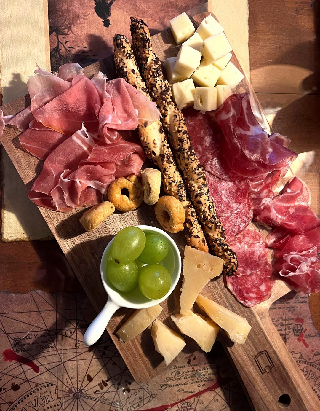 Tagliere visto dall'alto con prosciutto crudo, salame, formaggi, grissini ai semi, taralli e uva bianca, appoggiato su una carta nautica.
